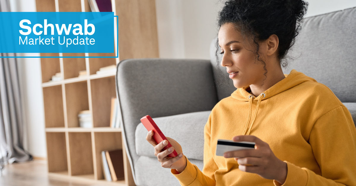 Schwab Market Update: A woman sits in front of her couch, holding a credit card and smartphone. 